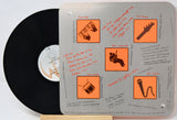 A vinyl record partially visible behind a square metal album cover insert with orange icons and handwritten notes. The record label shows the text 'A M.