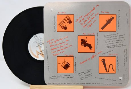 A vinyl record partially visible behind a square metal album cover insert with orange icons and handwritten notes. The record label shows the text 'A M.