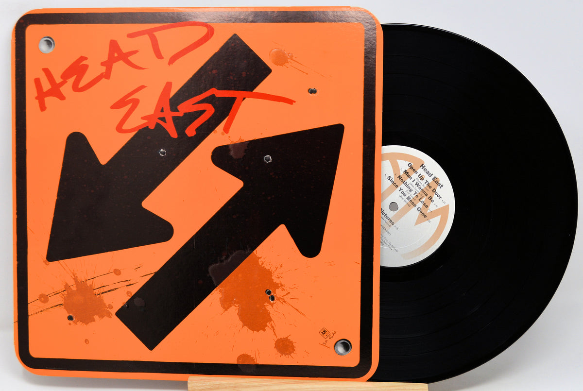 An orange square vinyl record sleeve or art print with black arrows, red graffiti-style text reading 'HEAD EAST,' and orange paint splatter details, shown next to a black vinyl record on a white background.
