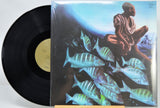 A vinyl record partially pulled from its illustrated album sleeve, showing a black record with a gold center label beside cover art of a seated figure above a school of fish.