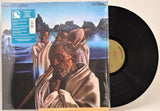A vinyl record album in its outer plastic shrink wrap, with the front cover art showing a painted scene of robed figures by a body of water and the black record partially visible behind it.