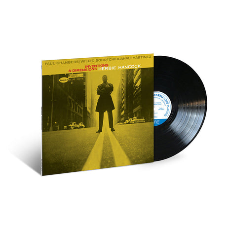 A vinyl record of 'Inventions & Dimensions' by Herbie Hancock with a yellow and black cover featuring a photograph of Herbie Hancock in the center, standing on a street corner.