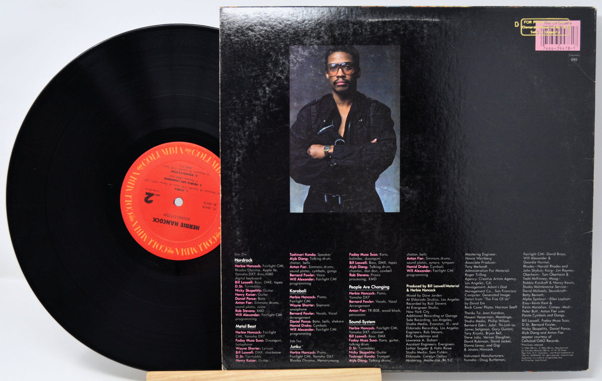 A vinyl record partially pulled from its black album cover, showing the back of the package with a portrait photo and track listings. The record label has a bright orange-red center label.