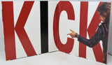 A large printed panel or book-like display featuring oversized red, white, and black letters spelling KICK, with a partial photo of a person in a dark jacket pointing toward the letter K. The product appears to be the printed panel itself rather than a lifestyle scene, so this is classified as Product/Packaging.