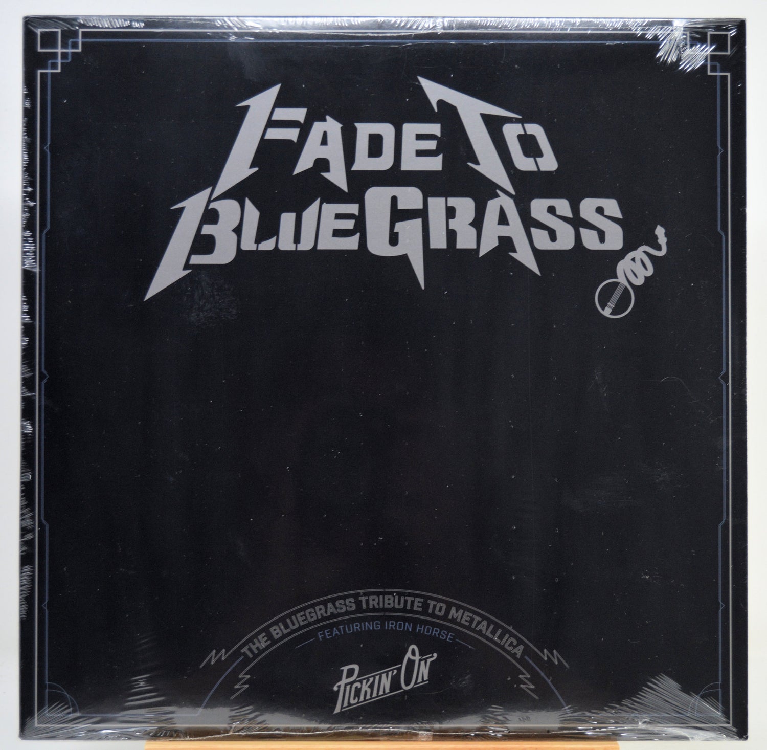 A square black album cover for 'Fade To Blue Grass' wrapped in clear plastic, featuring a white stylized logo and text, photographed against a white background.