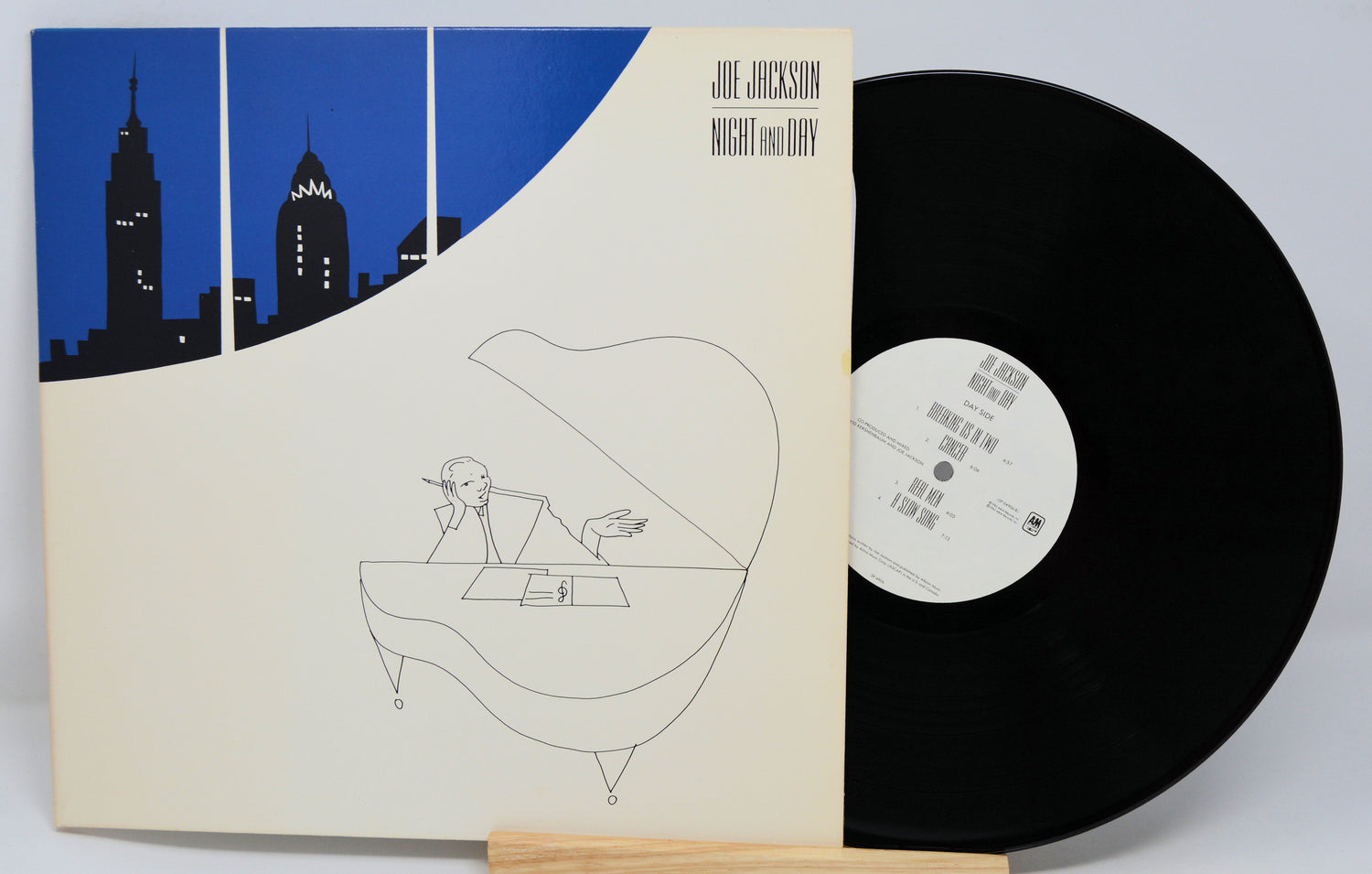 A vinyl record titled 'Night And Day' by Joe Jackson, featuring a cover illustration of a person at a piano with a city skyline in the background.