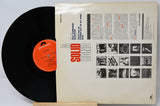 Vinyl record with its cover displaying text and black and white images on a light gray background