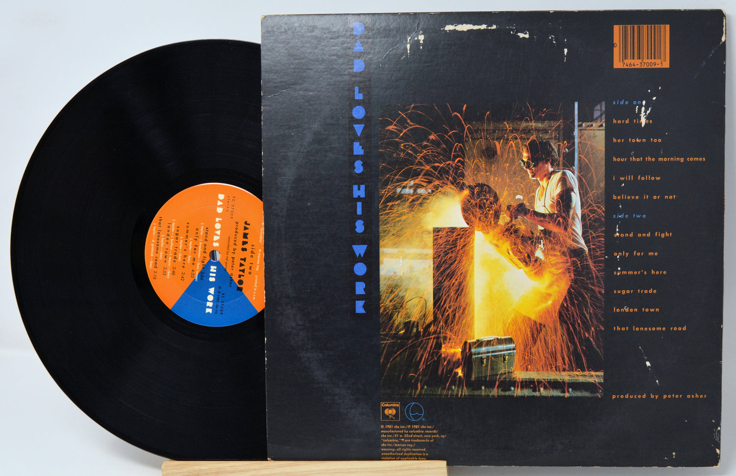 A vinyl record partially pulled from its sleeve, showing the black record, orange and blue center label, and the back cover artwork with a fiery welding scene and track listing.