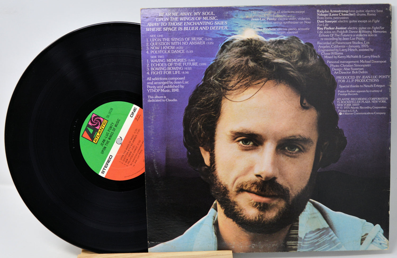 A vinyl record partially pulled from its sleeve, showing the black record with a green, orange, and white center label next to the printed album cover featuring a portrait of a bearded man against a purple background.