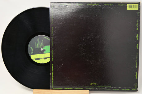 A vinyl record partially pulled from its sleeve, photographed on a light background. The record is black with a green-and-black center label, and the sleeve is predominantly black with thin green border text and a small green label in the top right corner.