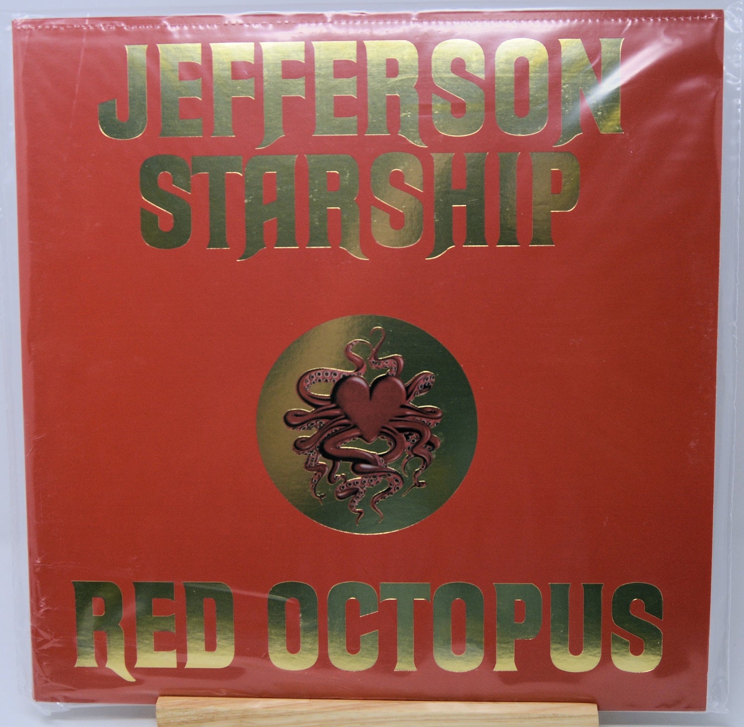 Jefferson Starship 'Red Octopus' vinyl record cover, featuring a textured red background with metallic gold lettering and a central gold medallion containing a red octopus and heart design. The record is sealed in clear plastic shrink-wrap and displayed on a wooden stand.