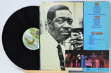 A vinyl record partially pulled from its album cover, showing the black record on the left and the printed cover art on the right. The cover features a black-and-white portrait of a man, a bright blue tracklist panel, and small performance photos at the bottom.