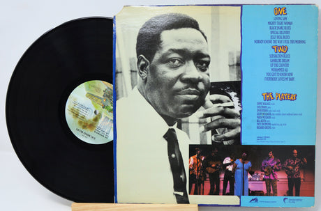 A vinyl record partially pulled from its album cover, showing the black record on the left and the printed cover art on the right. The cover features a black-and-white portrait of a man, a bright blue tracklist panel, and small performance photos at the bottom.