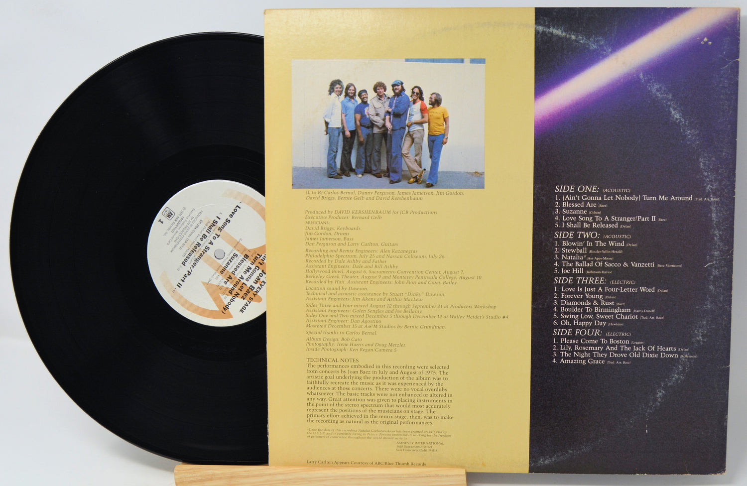A vinyl record partially pulled from its outer paper sleeve, showing the black record, printed tracklist on the right, and a pale yellow center label with a small band photo in the middle.