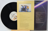 A vinyl record partially pulled from its outer paper sleeve, showing the black record, printed tracklist on the right, and a pale yellow center label with a small band photo in the middle.