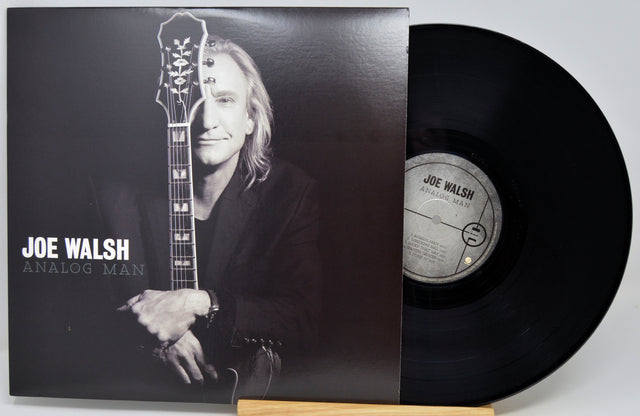 Joe Walsh 'Analog Man' vinyl record LP with its album cover sleeve and the black vinyl disc partially pulled out, displayed on a small wooden stand against a light gray background.
