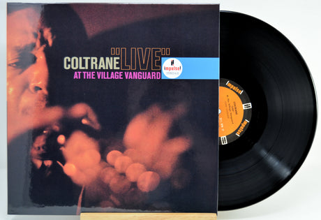 Coltrane 'Live At The Village Vanguard' vinyl record with the album cover standing beside the black LP disc on a light background.