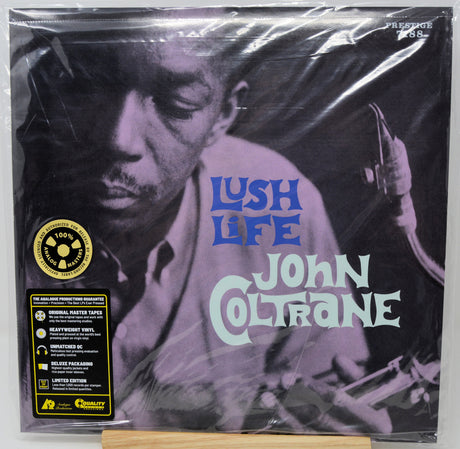 A sealed vinyl record of John Coltrane's 'Lush Life' on a wooden display stand. The album cover features a black and white portrait of the jazz musician, with 'Lush Life' in blue and 'John Coltrane' in light green text.