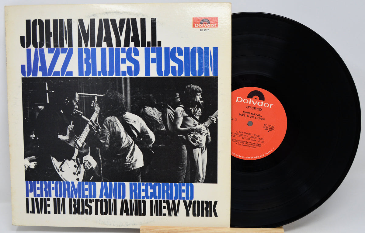 Vinyl record album for John Mayall's 'Jazz Blues Fusion' with the front cover standing upright and the black record partially pulled out to the right. The cover has a cream background with bold black and blue text and a black-and-white band photo, while the vinyl disc has a red-orange center label.