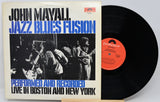 Vinyl record album for John Mayall's 'Jazz Blues Fusion' with the front cover standing upright and the black record partially pulled out to the right. The cover has a cream background with bold black and blue text and a black-and-white band photo, while the vinyl disc has a red-orange center label.
