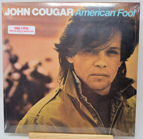 John Cougar 'American Fool' 12-inch vinyl LP record sleeve, sealed in plastic shrink wrap and displayed on a wooden stand. The cover features a portrait of the artist against a warm, sepia-toned background with the title in light blue text.