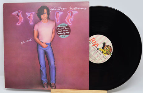 A vinyl record album shown with its printed cover partially covering the black record, photographed on a light gray background. The cover features a portrait of John Lennon against a pink-purple backdrop with stylized pink figures and the text 'John Lennon - Rebel Rebel'.