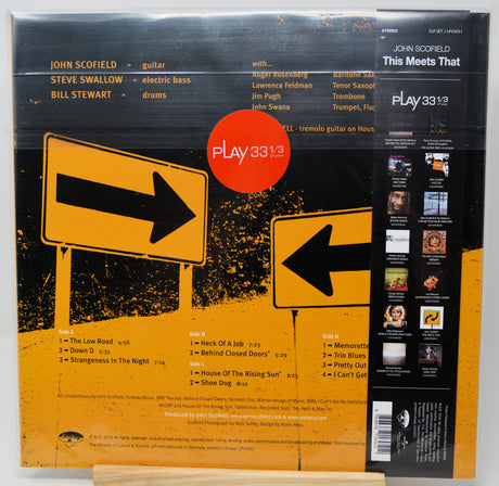 The back cover of a vinyl record for John Scofield's 'This Meets That' on Play 33&1/3. The design features a black background with a yellow-orange distressed texture, two road sign illustrations (one with a right arrow, one with a left arrow), and a central red circular logo containing the text 'PLAY 33&1/3'.