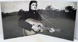 A black and white photographic print or poster depicting a man holding an acoustic guitar in an outdoor field, shown standing against a white background.