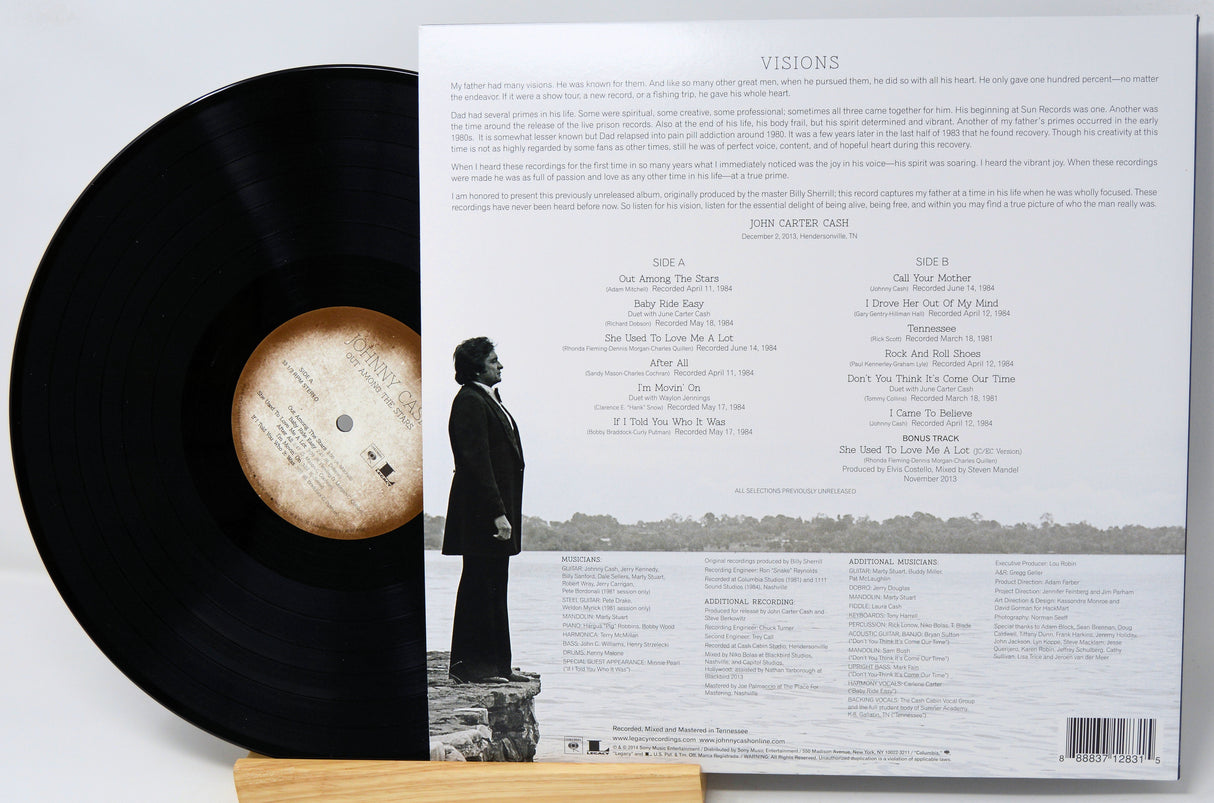 Vinyl record partially pulled from its open back cover, showing the black record, printed tracklist and liner notes, and a small printed photo of a person standing by water. The record rests on a small wooden stand against a light neutral background.