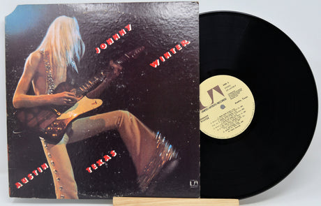 Johnny Winter Austin Texas vinyl record LP with its illustrated cover sleeve and the black vinyl disc partially pulled out, displayed on a small stand against a light gray background.