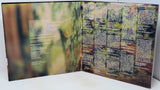 An open printed booklet or album-style book spread on a white surface, showing two printed pages with blurred forest imagery and dense text blocks. No person or real environment is present beyond the tabletop, so this is Product/Packaging rather than Lifestyle.
