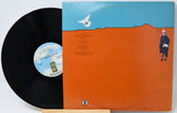 A vinyl record partially pulled from its sleeve, showing the black record and the back cover artwork. The sleeve has a bright orange lower half, a light blue upper band, a white dove illustration, and a small standing figure on the right.