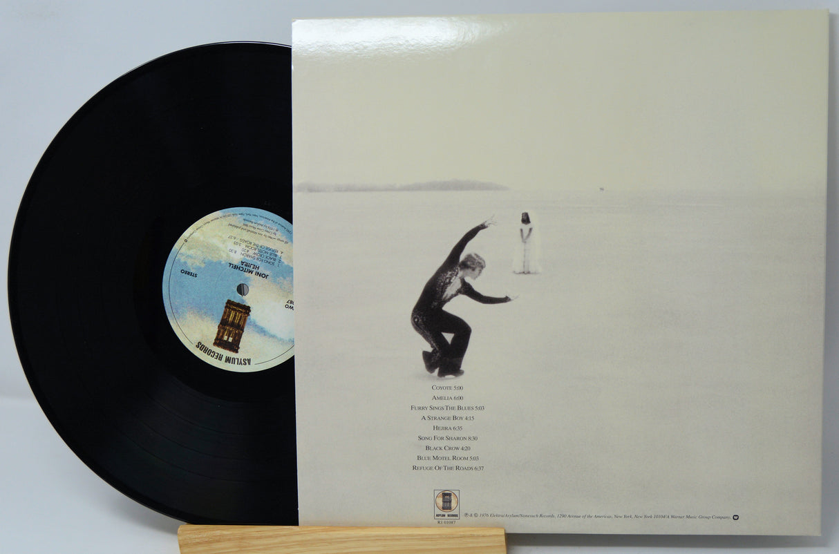 A vinyl record partially pulled from its cream-colored outer sleeve, displayed on a small wooden stand. The record is black with a blue and white center label, and the sleeve features a minimalist illustration of a figure in a vast white landscape.