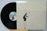 A vinyl record partially pulled from its cream-colored outer sleeve, displayed on a small wooden stand. The record is black with a blue and white center label, and the sleeve features a minimalist illustration of a figure in a vast white landscape.