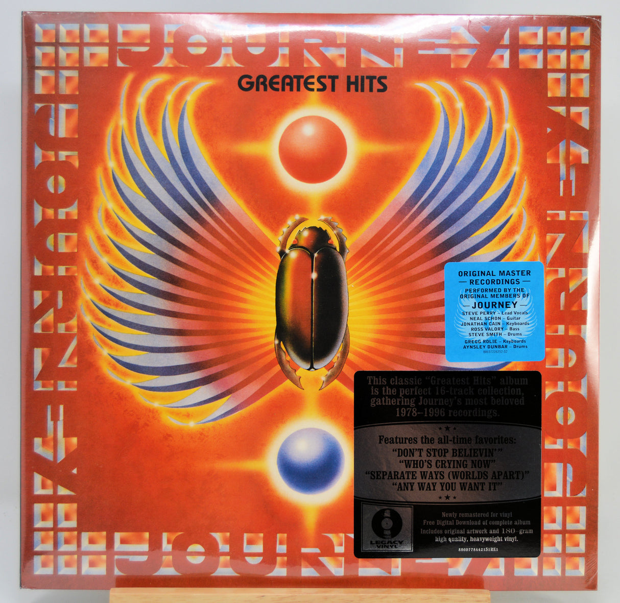 Journey 'Greatest Hits' vinyl record album cover, showing a glowing orange and red Egyptian-style winged emblem with a beetle motif, a blue original master sticker, and a black tracklist sticker on a white background.