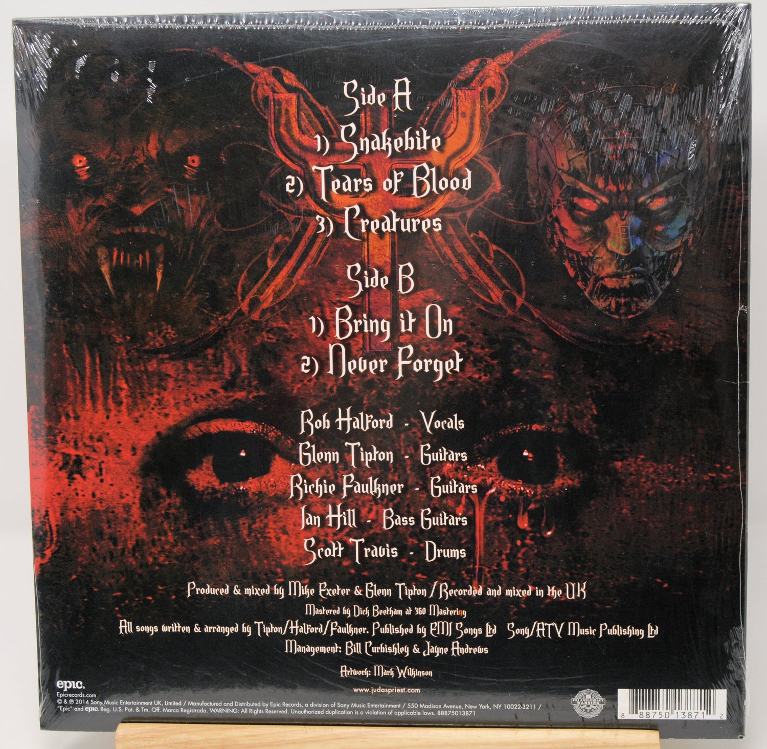 The back cover of a vinyl record for the band Quakehouse's album 'Tears of Blood', sealed in shrink wrap and displayed on a wooden stand. The artwork is dark and horror-themed, featuring red demonic faces, a large eye, and white text listing the tracklist.