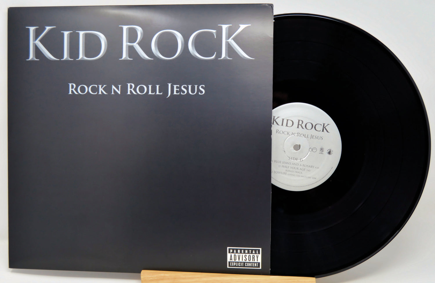 A vinyl record album for 'Rock N Roll Jesus' by Kid Rock, showing the black cover with silver text and a black vinyl record partially pulled out, displayed on a wooden stand against a light background.