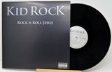 A vinyl record album for 'Rock N Roll Jesus' by Kid Rock, showing the black cover with silver text and a black vinyl record partially pulled out, displayed on a wooden stand against a light background.