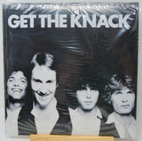 A vinyl record sleeve for the band 'Get The Knack' wrapped in clear plastic, displayed on a small wooden stand against a white background.