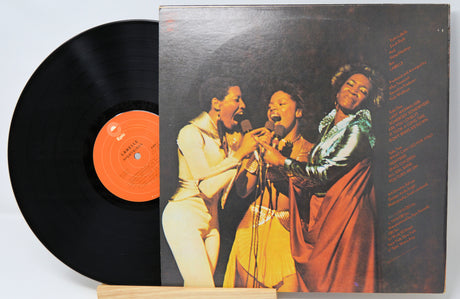 Vinyl record with a colorful cover featuring three people singing, on a light gray background.