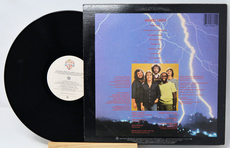 A vinyl record partially pulled from its back cover sleeve, showing the black record on the left and the printed back cover on the right. The sleeve features a blue lightning scene with a band photo in the center and track listing text.