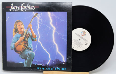 Larry Carlton's 'Strikes Twice' vinyl record LP with its illustrated album cover sleeve and the black vinyl record partially pulled out, displayed on a small wooden stand against a light gray background.