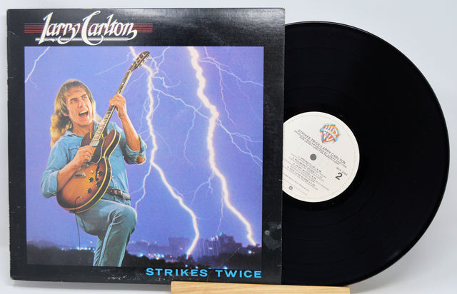 Larry Carlton's 'Strikes Twice' vinyl record LP with its illustrated album cover sleeve and the black vinyl record partially pulled out, displayed on a small wooden stand against a light gray background.