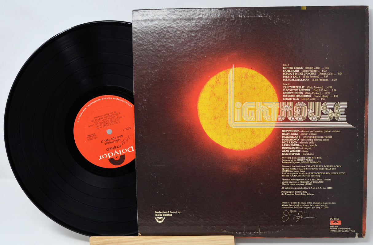 A vinyl record partially pulled from its album cover, showing the black record with an orange center label and the back cover featuring a large glowing orange sun and the word 'Lighthouse' in white.
