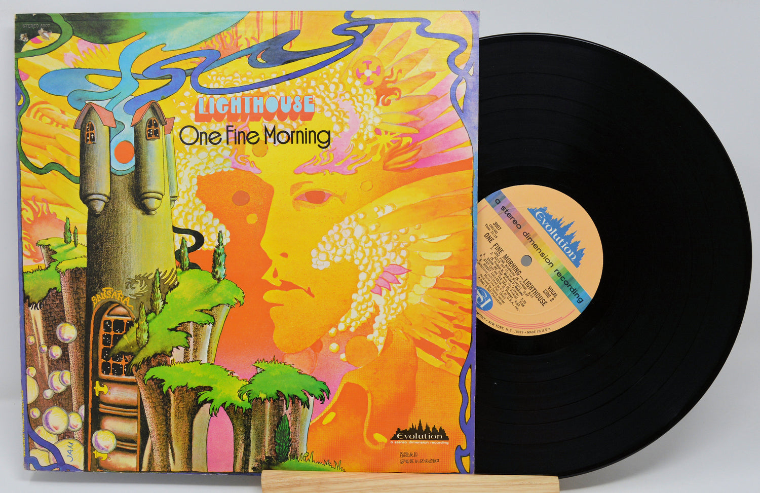 A vinyl record and its illustrated album cover for Lighthouse's 'One Fine Morning' displayed on a small wooden stand against a light gray background.