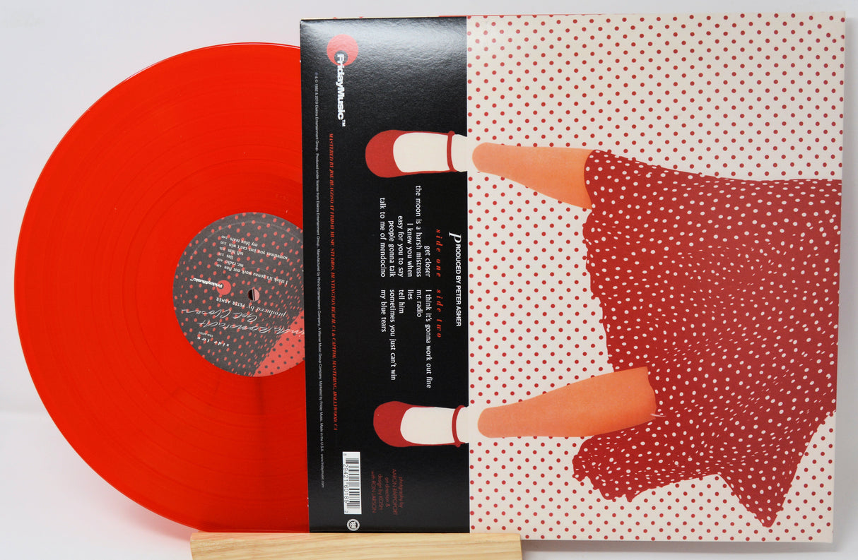 A vinyl record partially pulled from its illustrated album cover, showing a translucent bright red record and matching cover art with a stylized figure in a red polka-dot dress on a white background.
