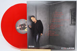A red vinyl record partially pulled from its sleeve, showing the back cover with tracklist text and a printed portrait of a man in a dark outfit against a gray wall.