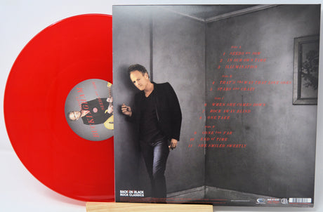 A red vinyl record partially pulled from its sleeve, showing the back cover with tracklist text and a printed portrait of a man in a dark outfit against a gray wall.