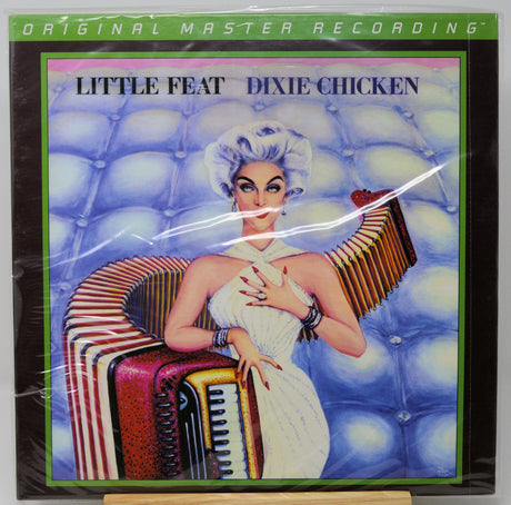 Vinyl record sleeve for 'Little Feat Dixie Chicken' on a black background, wrapped in clear plastic. The cover art shows a retro-style illustrated woman in a white dress seated on an accordion against a blue tufted backdrop, with a green border and 'Original Master Recording' text at the top.