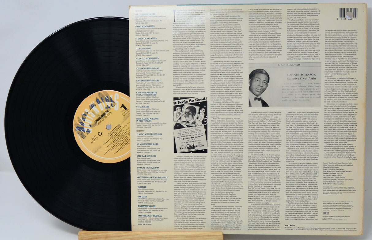 A vinyl record partially pulled from its back cover sleeve, displayed on a small wooden stand against a light gray background. The black record has a tan label, and the sleeve is cream-colored with dense text and a small black-and-white portrait.